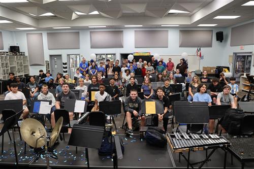 A group of band students sit with instruments and adults stand behind them
