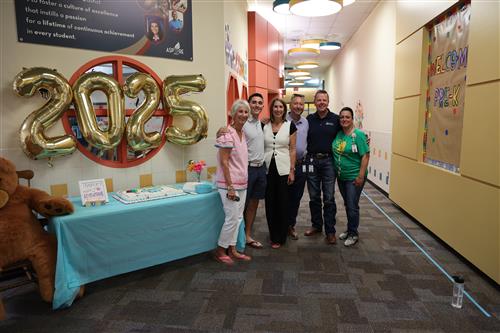 A group of people stand next to a table with cake on it and balloons spelling out the words 20 25