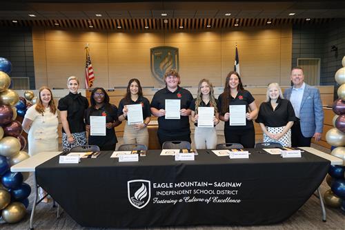 Students stand behind a table holding certificates and flanked by adults