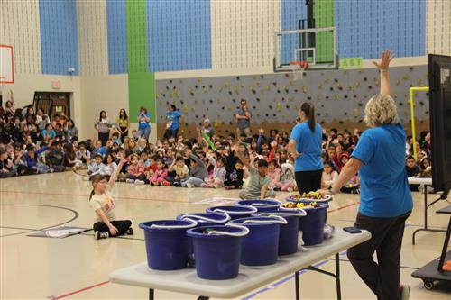 A woman holds up a hand in front of a large audience in a school gym
