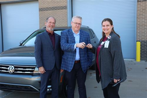 Two men hand a set of keys to a woman with a car behind them