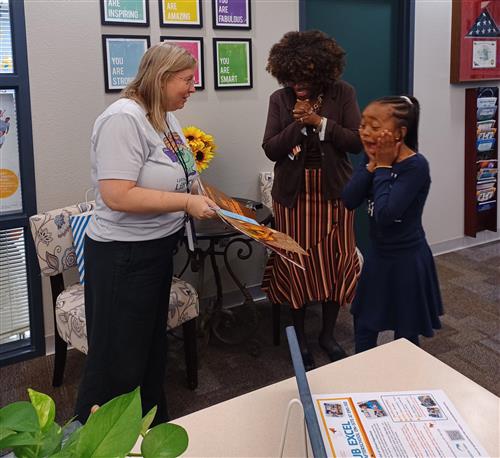 A girl screams while her mom smiles and a woman hands her a book